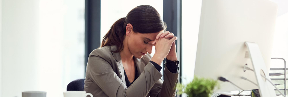A woman is seen sitting at her work table, looking worried, with her hands folded and resting on her forehead.
